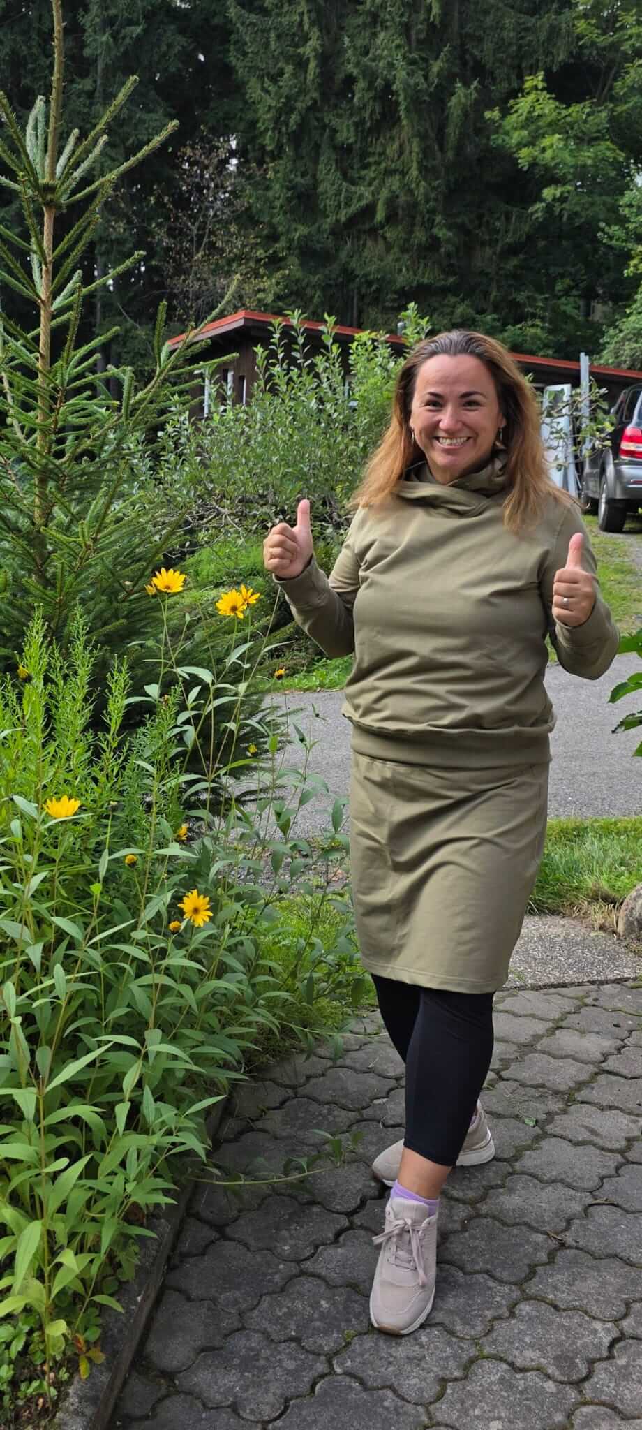 Žena s úsměvem v khaki mikině s kapucí, stojící v zahradě mezi květinami a zelení.